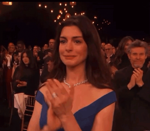 Anne Hathaway claps with teary eyes at a formal event.