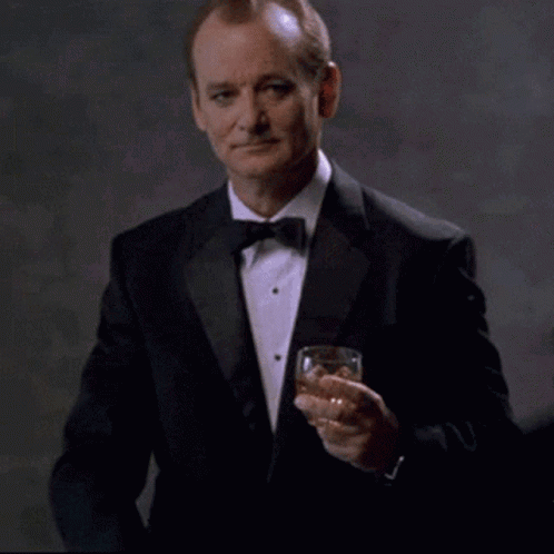 A man in a tuxedo holds up a drink with a restrained, approving expression.