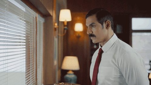 a man with a mustache in a white shirt and red tie looks out a window