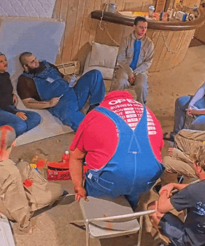 a group of men are sitting around a man wearing overalls that says open business hours