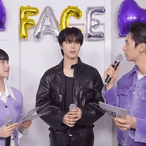 a man in a black leather jacket holds a microphone in front of balloons that say face