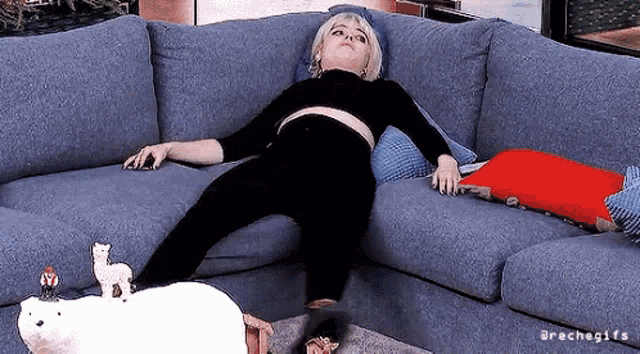 A person lies spread out across a couch surrounded by laundry and cushions.