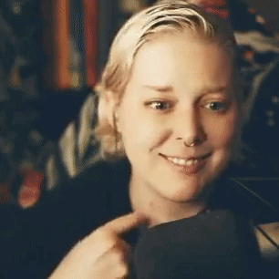A blonde person pulls at their shirt collar with an uncomfortable smile.