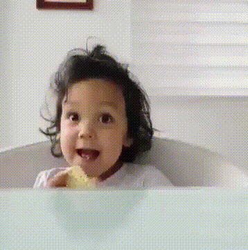 A toddler in a bathtub smiles and holds up a thumb.