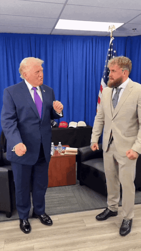 a man in a suit stands next to a man in a tan suit in front of an american flag