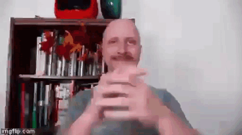 A bald man claps quickly in front of a bookcase.