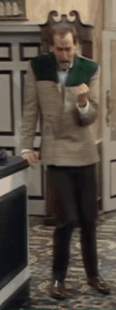a man in a tan jacket and black pants is standing in a room with a door in the background .