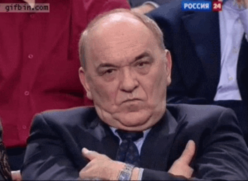 An older man sits with folded arms and a stern frown.