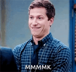 A man in a blue plaid shirt makes an uncomfortable closed-mouth smile.