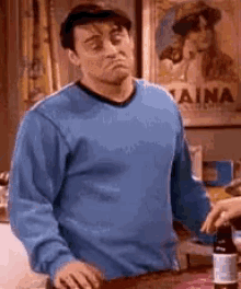 A man in a blue sweater makes a dismissive shrugging expression indoors.