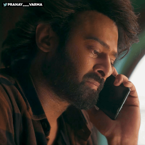 Prabhas holds a phone and looks sadly to the side in a dimly lit scene.