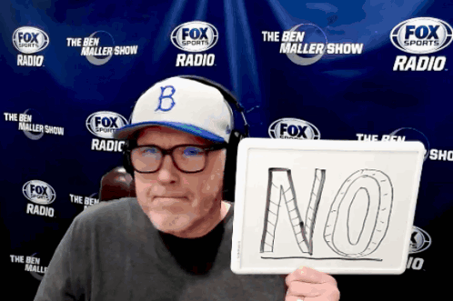 A man in a cap holds up a hand-drawn “NO” sign at a radio backdrop.