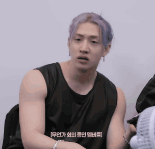 a man with purple hair is wearing a black tank top and sitting next to a fan