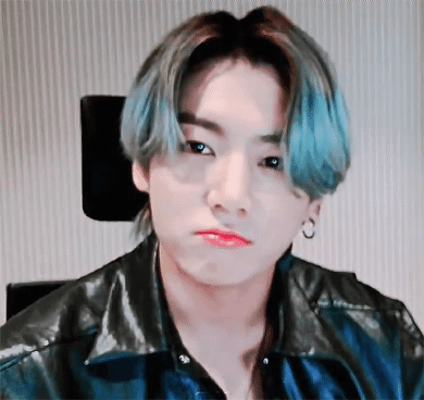 A man with teal-streaked hair pouts while seated in a black leather jacket.