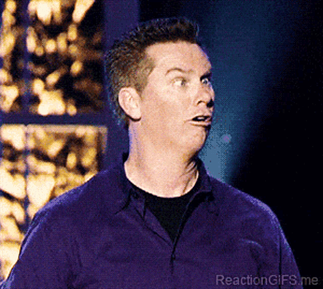 A comedian in a purple shirt makes a surprised, open-mouthed reaction.