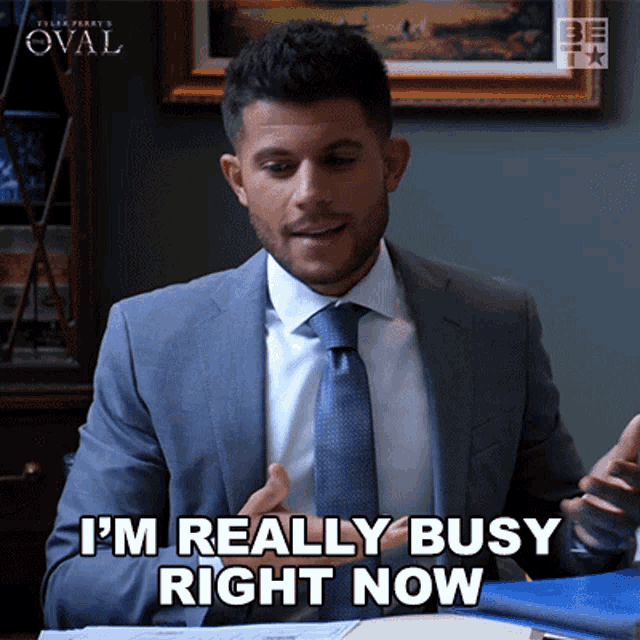 a man in a suit and tie is sitting at a desk and says i 'm really busy right now