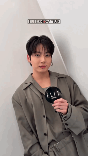 Jungkook in a gray outfit holds a microphone and poses indoors beneath small text.