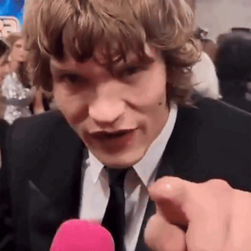a man in a suit and tie is pointing at the camera while talking into a pink microphone .