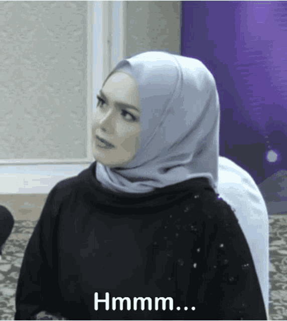 a woman wearing a hijab says " hmm " in front of a purple wall
