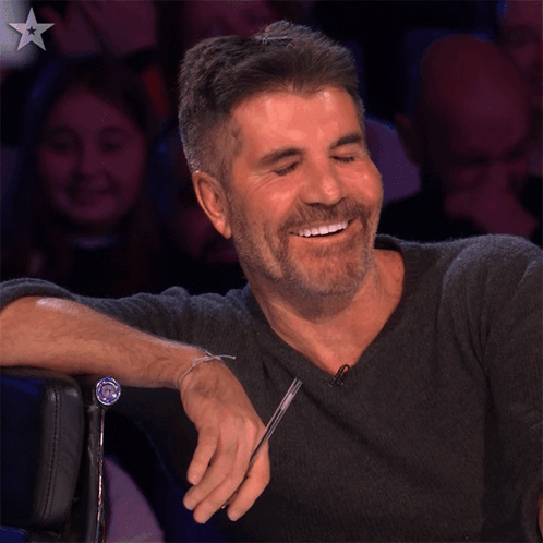 Simon Cowell laughs with his hand on his chest in a studio audience setting.