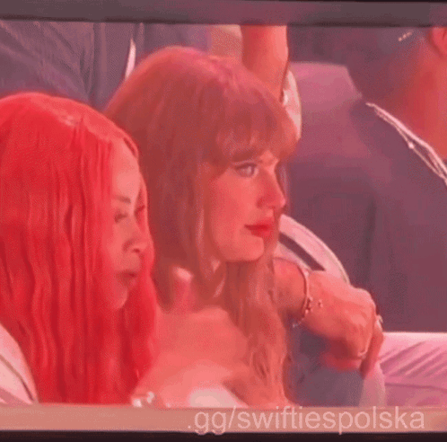 Taylor Swift watches from a crowded event, lit in red tones.