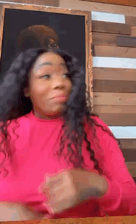 a woman in a pink shirt is sitting at a table with her hands folded in front of a wooden wall .