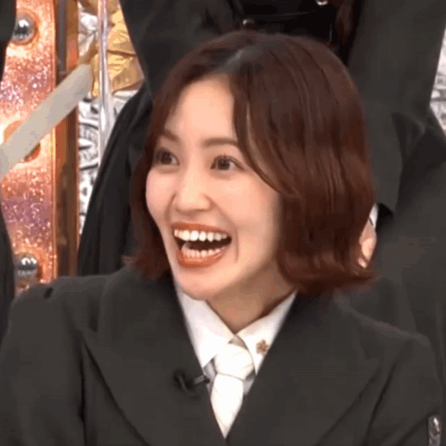 A woman in a black suit smiles broadly with delighted surprise.