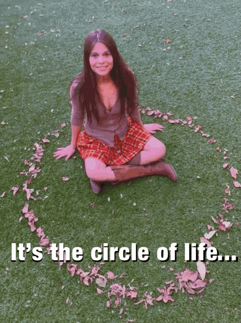 a woman in a plaid skirt sits on the grass with her arms outstretched and a circle of leaves around her knee