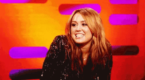 Miley Cyrus gives a hesitant smile on a brightly lit talk-show set.
