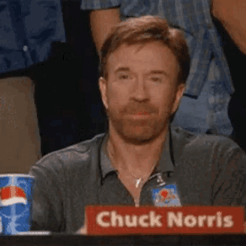 Chuck Norris sits holding a sign that reads “Chuck Norris” while giving a thumbs-up.