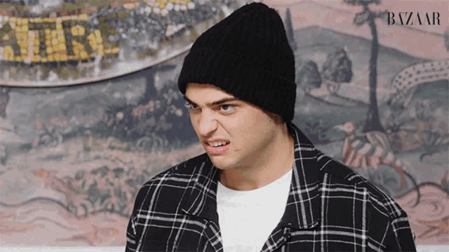 a man wearing a plaid shirt and a black beanie stands in front of a harper 's bazaar advertisement
