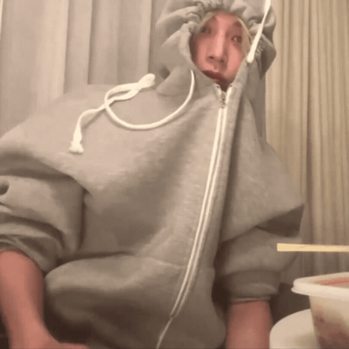A person in a gray hooded jacket pulls the zipper up over their face at a table.