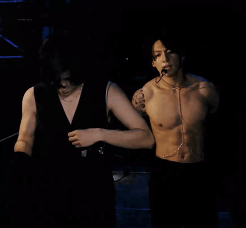 A shirtless man wearing chains stands in a dark performance scene.