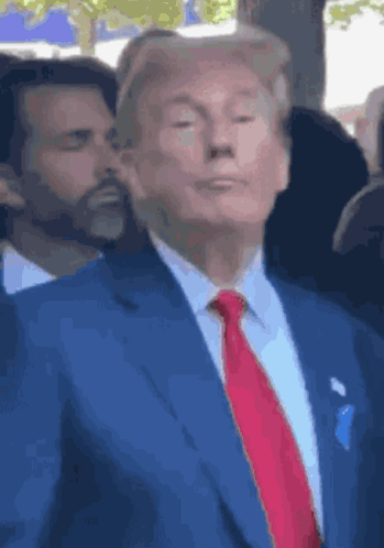 Donald Trump stands among people in a blue suit, looking forward.