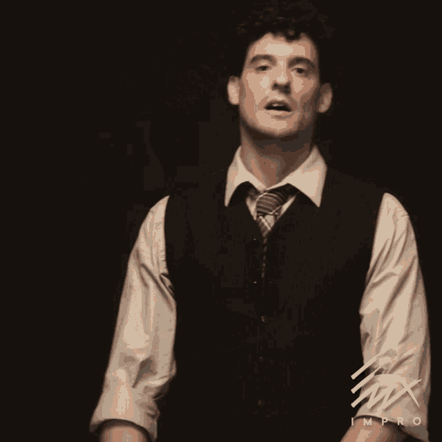 A man in a vest appears against a dark background with a downcast, uncertain expression.