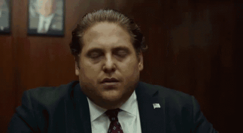 A suited man sits with his eyes closed, looking stressed and resigned.