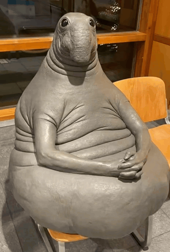 A bizarre seated creature with a birdlike head folds its arms in a restaurant.