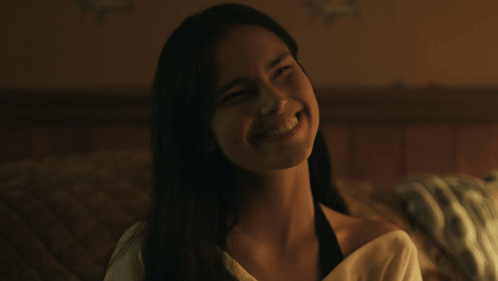 A woman smiles eerily while seated in a dim bedroom.