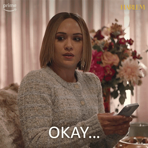 a woman in a grey sweater is holding a cell phone and says okay