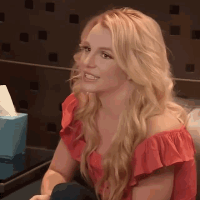 a blonde woman in a red off the shoulder top is sitting in front of a blue box of tissues .