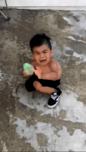 A child stands in water holding a green balloon and looks distressed.