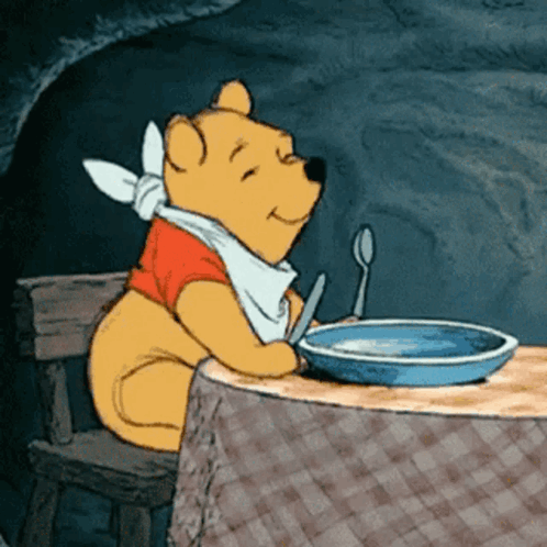 Winnie the Pooh waits at a table with an empty bowl and spoon.