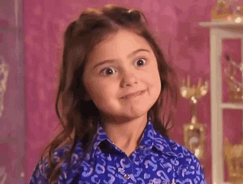 A young girl gives a wide, eager grin in a pink room.