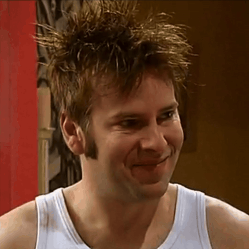 A man in a white tank top gives a reluctant, awkward smile.