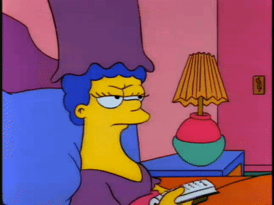 Marge Simpson sits by a lamp with a weary sideways look.
