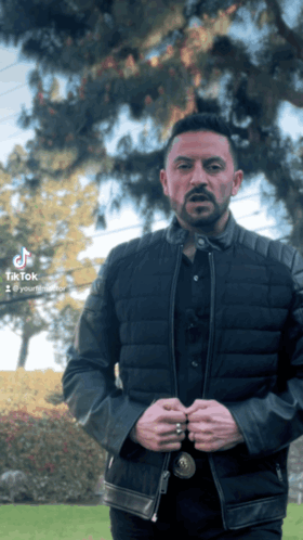 a man in a black jacket is standing in front of a tree with tik tok written on the bottom right
