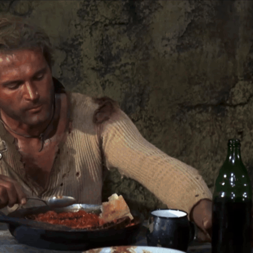 A rugged man eats beans and bread at a table.