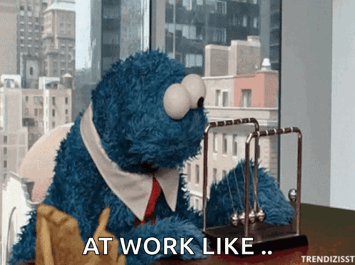 Cookie Monster sits at a home-office desk with the caption “AT WORK LIKE...”