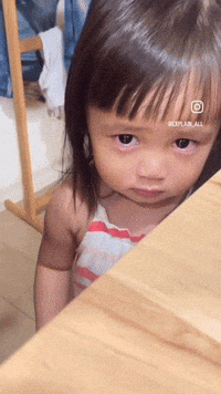A little girl peers up from beside a table with watery eyes.
