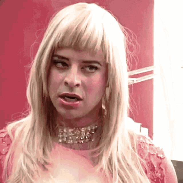 A person in a blonde wig stares forward, visibly offended and angry.